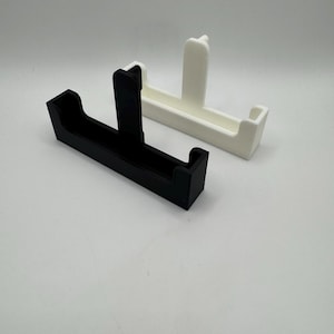 May include: Two 3D-printed holders, one black and one white, are displayed against a plain background. Each holder has a rectangular base with a vertical support in the center. The white holder is positioned behind the black one.