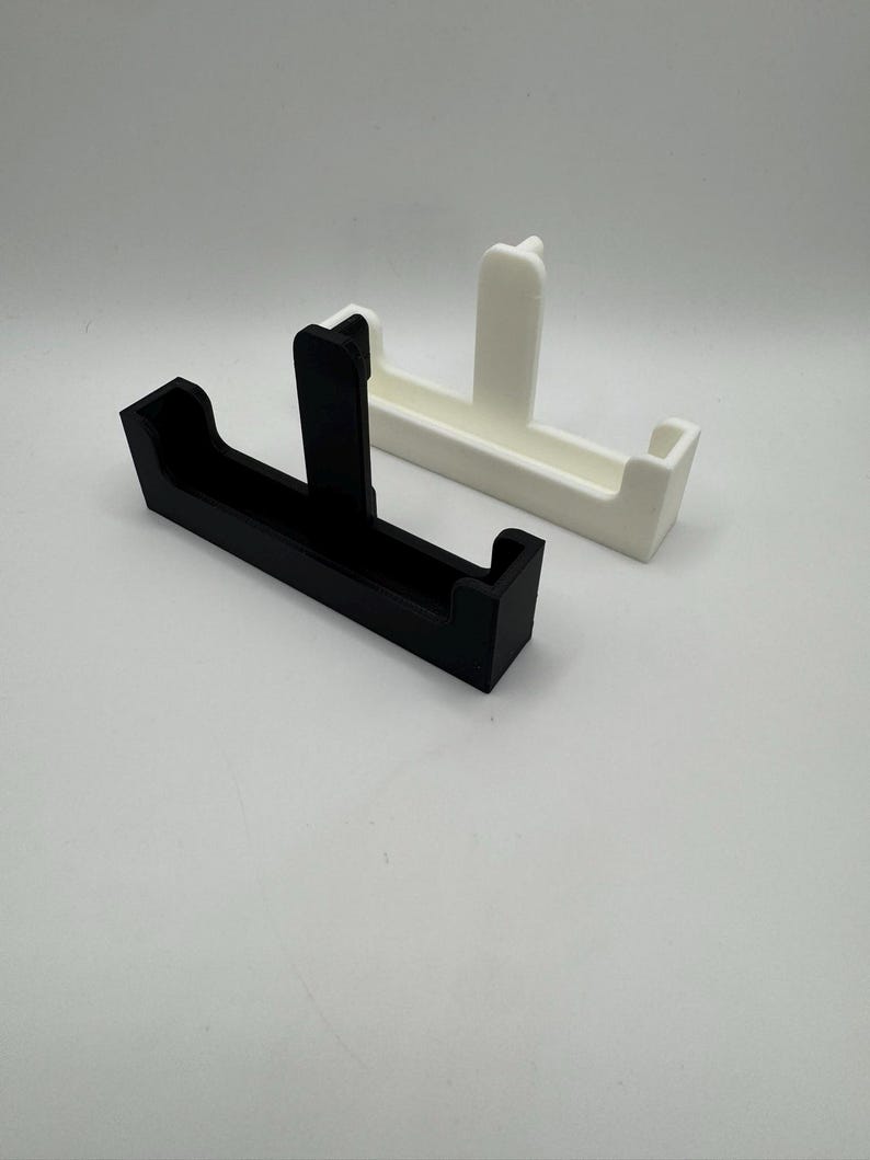 May include: Two 3D-printed holders, one black and one white, are displayed against a plain background. Each holder has a rectangular base with a vertical support in the center. The white holder is positioned behind the black one.