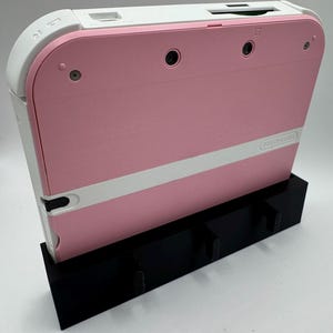 May include: A pink and white Nintendo 2DS handheld game console is displayed in a black stand. The console has a pink body with a white stripe and white top. The stand has three slots for game cartridges.