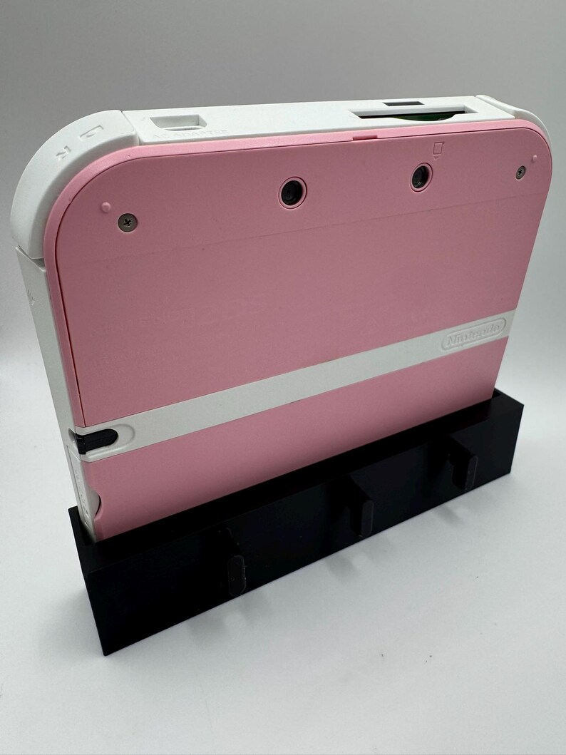 May include: A pink and white Nintendo 2DS handheld game console is displayed in a black stand. The console has a pink body with a white stripe and white top. The stand has three slots for game cartridges.