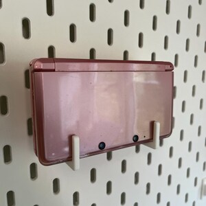 Puede incluir: Una consola de juegos port&aacute;til Nintendo 3DS de color oro rosa montada en una placa perforada blanca con soportes blancos. La consola est&aacute; cerrada, mostrando su superficie lisa y reflectante y dos peque&ntilde;as lentes negras.