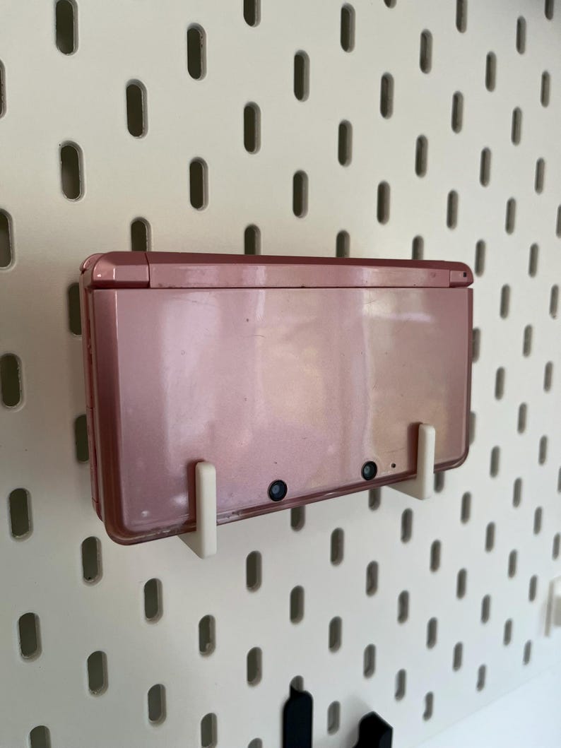 Puede incluir: Una consola de juegos port&aacute;til Nintendo 3DS de color oro rosa montada en una placa perforada blanca con soportes blancos. La consola est&aacute; cerrada, mostrando su superficie lisa y reflectante y dos peque&ntilde;as lentes negras.