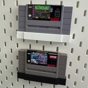 K&ouml;nnte beinhalten: Zwei Super Nintendo-Spielmodule, "PGA Tour Golf" und "Street Fighter II", werden auf einer wei&szlig;en Lochwand pr&auml;sentiert. Das Modul "PGA Tour Golf" hat ein gr&uuml;nes und lila Etikett. Das Modul "Street Fighter II" hat ein farbenfrohes Etikett mit einer Kampfszene.