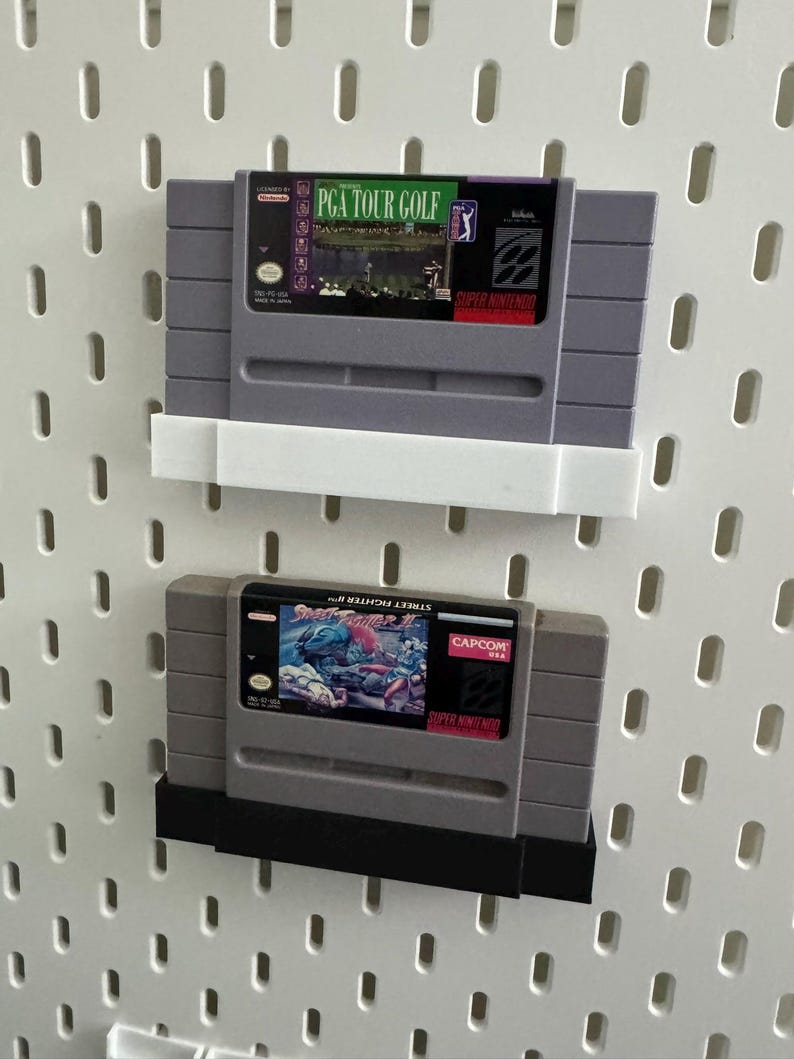K&ouml;nnte beinhalten: Zwei Super Nintendo-Spielmodule, "PGA Tour Golf" und "Street Fighter II", werden auf einer wei&szlig;en Lochwand pr&auml;sentiert. Das Modul "PGA Tour Golf" hat ein gr&uuml;nes und lila Etikett. Das Modul "Street Fighter II" hat ein farbenfrohes Etikett mit einer Kampfszene.
