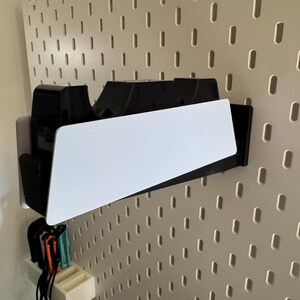 K&ouml;nnte beinhalten: Ein schwarz-wei&szlig;er Wandorganizer mit einem wei&szlig;en, abgewinkelten Regal. Der Organizer ist an einer Lochwand befestigt. Darunter befinden sich mehrere farbige Gegenst&auml;nde.