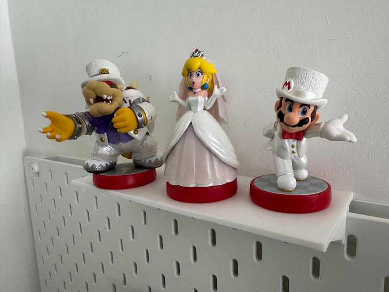 Puede incluir: Tres figuras blancas de boda de Mario, Peach y Bowser. Mario lleva un esmoquin blanco y un sombrero de copa con una 'M' roja. Peach lleva un vestido de novia blanco con velo y tiara. Bowser lleva un esmoquin blanco y un sombrero de copa con una banda morada. Las figuras est&aacute;n en un estante blanco.