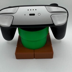 K&ouml;nnte beinhalten: Ein schwarzer und silberner Videospiel-Controller, der auf einem gr&uuml;nen Rohr und einem braunen Blockst&auml;nder ruht. Der Controller hat silberne Tasten und schwarze Griffe. Der St&auml;nder ist eine Nachbildung eines klassischen Spiellevels.