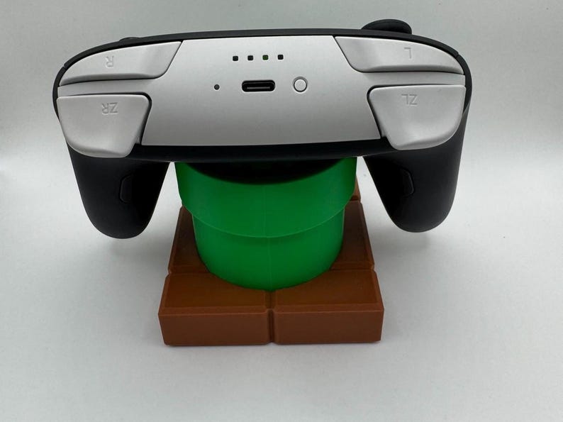 K&ouml;nnte beinhalten: Ein schwarzer und silberner Videospiel-Controller, der auf einem gr&uuml;nen Rohr und einem braunen Blockst&auml;nder ruht. Der Controller hat silberne Tasten und schwarze Griffe. Der St&auml;nder ist eine Nachbildung eines klassischen Spiellevels.