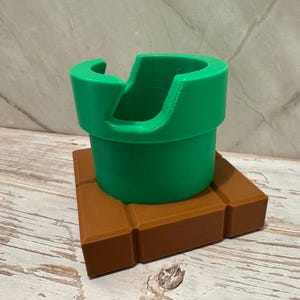 May include: A green pipe-shaped object with a curved opening, sitting on a brown brick-like base. The design is reminiscent of a video game theme. The pipe is approximately 10 cm tall.