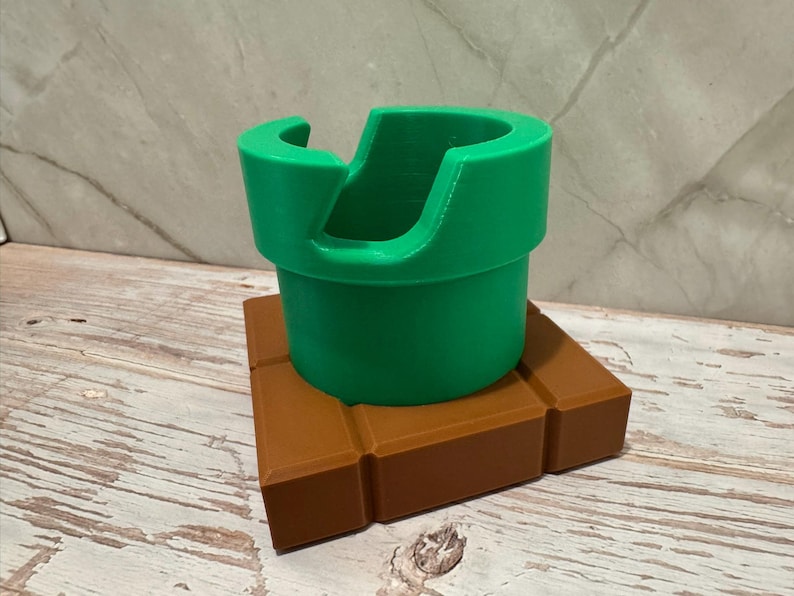 May include: A green pipe-shaped object with a curved opening, sitting on a brown brick-like base. The design is reminiscent of a video game theme. The pipe is approximately 10 cm tall.