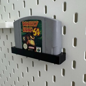May include: A gray Donkey Kong 64 video game cartridge with a colorful label featuring Donkey Kong. The cartridge is displayed in a black shelf against a white pegboard wall. The game is for the Nintendo 64.