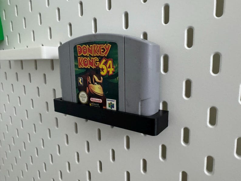 May include: A gray Donkey Kong 64 video game cartridge with a colorful label featuring Donkey Kong. The cartridge is displayed in a black shelf against a white pegboard wall. The game is for the Nintendo 64.