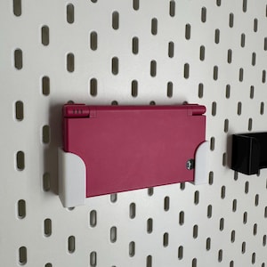 Puede incluir: Una consola de juegos portátil Nintendo DS magenta montada en una pared perforada blanca con soportes blancos. La pared perforada tiene agujeros ovalados espaciados uniformemente. También hay un soporte negro montado.