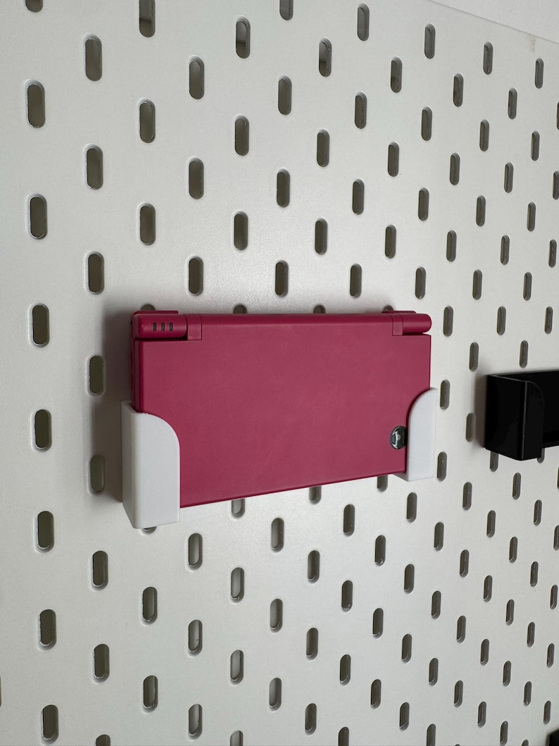 K&ouml;nnte beinhalten: Eine magenta Nintendo DS Handheld-Spielkonsole, montiert auf einer wei&szlig;en Lochwand mit wei&szlig;en Halterungen. Die Lochwand hat gleichm&auml;&szlig;ig verteilte ovale L&ouml;cher. Eine schwarze Halterung ist ebenfalls montiert.