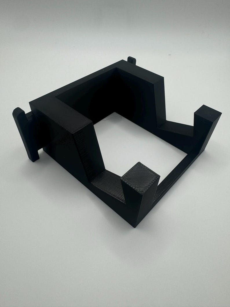 K&ouml;nnte beinhalten: Ein schwarzer 3D-gedruckter St&auml;nder f&uuml;r ein Ger&auml;t mit quadratischer Basis und einer erh&ouml;hten Plattform.