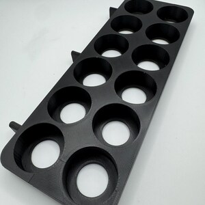 May include: A black, rectangular shot glass holder with 12 circular openings. The holder is made of a textured material and has two small, angled supports on one side. The openings are evenly spaced and appear to be designed to hold shot glasses.