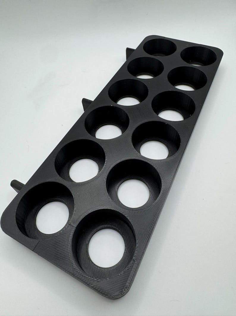 May include: A black, rectangular shot glass holder with 12 circular openings. The holder is made of a textured material and has two small, angled supports on one side. The openings are evenly spaced and appear to be designed to hold shot glasses.