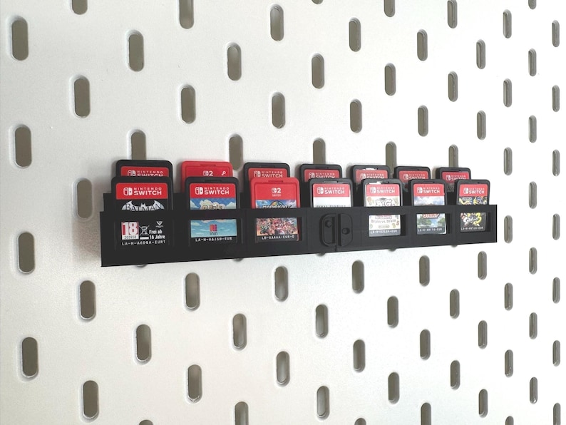 K&ouml;nnte beinhalten: Ein schwarzer, an der Wand befestigter Spielkassettenhalter, gef&uuml;llt mit Nintendo Switch-Spielkassetten. Die Kassetten sind rot und schwarz. Der Halter ist an einer wei&szlig;en Lochwand befestigt.