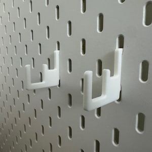 May include: White pegboard with two white hooks. The pegboard has a grid of evenly spaced, oblong holes. The hooks are designed to fit into the holes, providing a storage or display solution. The hooks are in a U-shape.