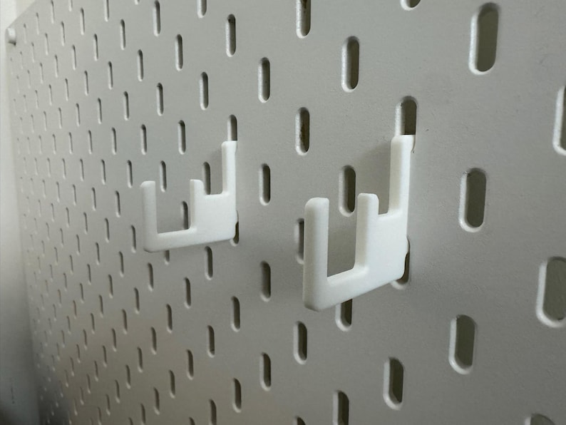 May include: White pegboard with two white hooks. The pegboard has a grid of evenly spaced, oblong holes. The hooks are designed to fit into the holes, providing a storage or display solution. The hooks are in a U-shape.