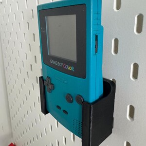 May include: A teal Game Boy Color handheld video game console is mounted on a white pegboard wall using a black 3D printed wall mount.