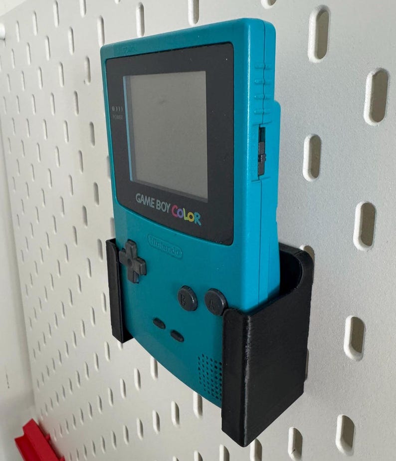 May include: A teal Game Boy Color handheld video game console is mounted on a white pegboard wall using a black 3D printed wall mount.