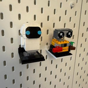 May include: Two small white pegboard shelves with a white and black robot and a yellow and black robot on them. The white robot has blue eyes and the yellow robot has a red toolbox and a plant.