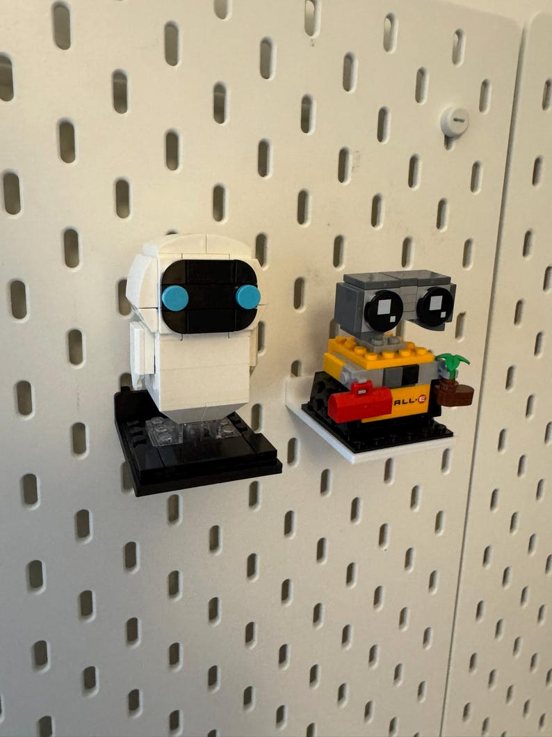 May include: Two small white pegboard shelves with a white and black robot and a yellow and black robot on them. The white robot has blue eyes and the yellow robot has a red toolbox and a plant.
