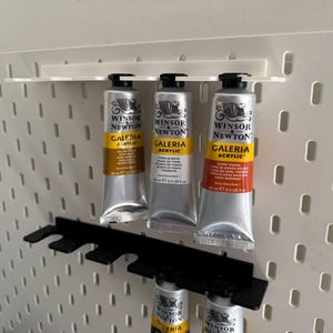 May include: Three tubes of Winsor & Newton Galeria acrylic paint in silver tubes with yellow and orange labels, displayed on a white pegboard with black holders. The tubes are labelled with the paint colour and brand.