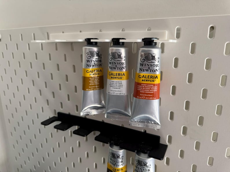May include: Three tubes of Winsor & Newton Galeria acrylic paint in silver tubes with yellow and orange labels, displayed on a white pegboard with black holders. The tubes are labelled with the paint colour and brand.