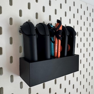 以下が含まれることがあります： 黒い収納オーガナイザー。黒いコントローラーとオレンジと青のゲームコントローラーを収納する複数のコンパートメントがあります。 オーガナイザーは、楕円形の穴のある白いペグボードに取り付けられています。