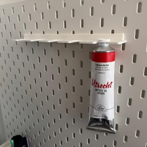 May include: A white pegboard wall organizer featuring a horizontal rack with slots for hanging items. A tube of Utrecht Cadmium Red Hue artists' oil paint is suspended from one of the slots by its cap. The pegboard has a grid of vertical oval holes.