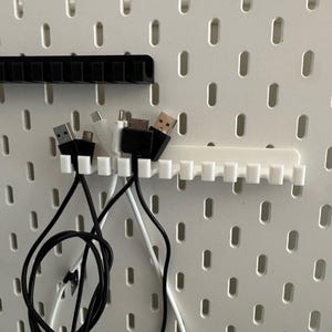 May include: White and black cable organizers with multiple slots, holding various USB cables. The organizers are mounted on a white pegboard, providing a tidy solution for cable management.