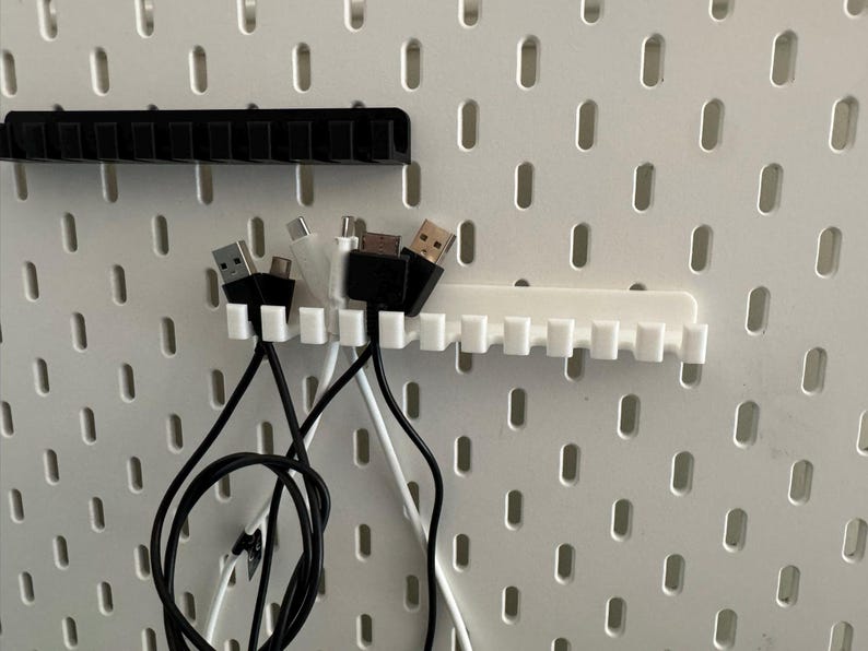 May include: White and black cable organizers with multiple slots, holding various USB cables. The organizers are mounted on a white pegboard, providing a tidy solution for cable management.