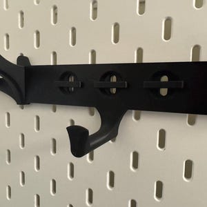 May include: Black pegboard organizer with multiple hooks and circular cutouts. The organizer is designed to attach to a pegboard and provides a storage solution. The hooks are curved and angled, suitable for hanging items.