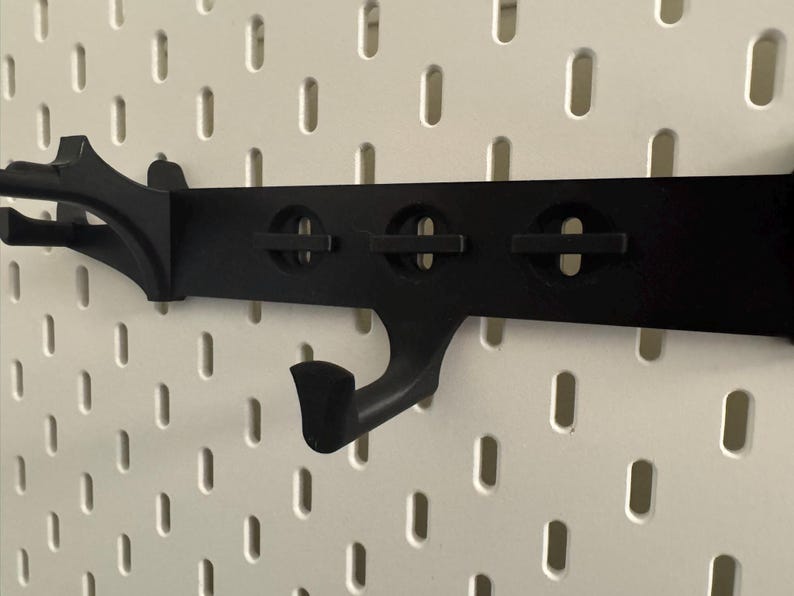 May include: Black pegboard organizer with multiple hooks and circular cutouts. The organizer is designed to attach to a pegboard and provides a storage solution. The hooks are curved and angled, suitable for hanging items.