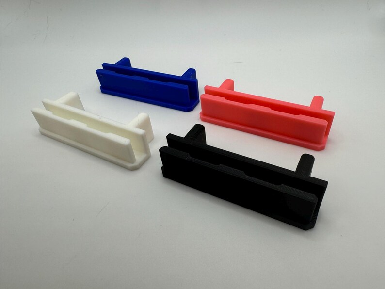 Pode incluir: Quatro suportes de prateleira de pl&aacute;stico em diferentes cores: branco, azul, rosa e preto. Cada suporte tem duas ranhuras e uma borda elevada na parte superior.