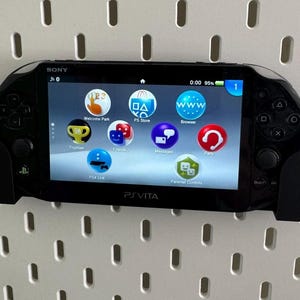Op de afbeelding: Een zwarte Sony PlayStation Vita handheld-gameconsole met een wit scherm waarop het startmenu wordt weergegeven. De console heeft een zwarte en grijze controller aan de rechterkant en een zwarte en grijze controller aan de linkerkant. Het scherm toont pictogrammen voor Welcome Park, PS Store, Browser, Berichten, Party en Ouderlijk toezicht.