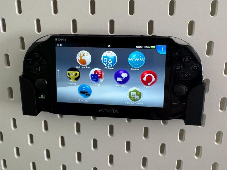 Op de afbeelding: Een zwarte Sony PlayStation Vita handheld-gameconsole met een wit scherm waarop het startmenu wordt weergegeven. De console heeft een zwarte en grijze controller aan de rechterkant en een zwarte en grijze controller aan de linkerkant. Het scherm toont pictogrammen voor Welcome Park, PS Store, Browser, Berichten, Party en Ouderlijk toezicht.