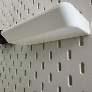 Puede incluir: Una estanter&iacute;a de pl&aacute;stico blanco con bordes redondeados est&aacute; montada en una pared de tablero de clavijas blanco. La estanter&iacute;a est&aacute; vac&iacute;a.