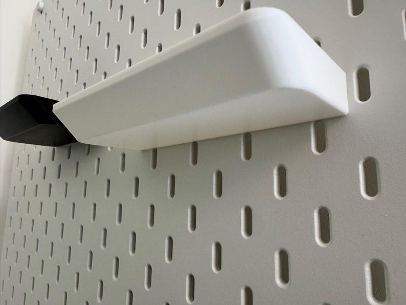 Puede incluir: Una estanter&iacute;a de pl&aacute;stico blanco con bordes redondeados est&aacute; montada en una pared de tablero de clavijas blanco. La estanter&iacute;a est&aacute; vac&iacute;a.