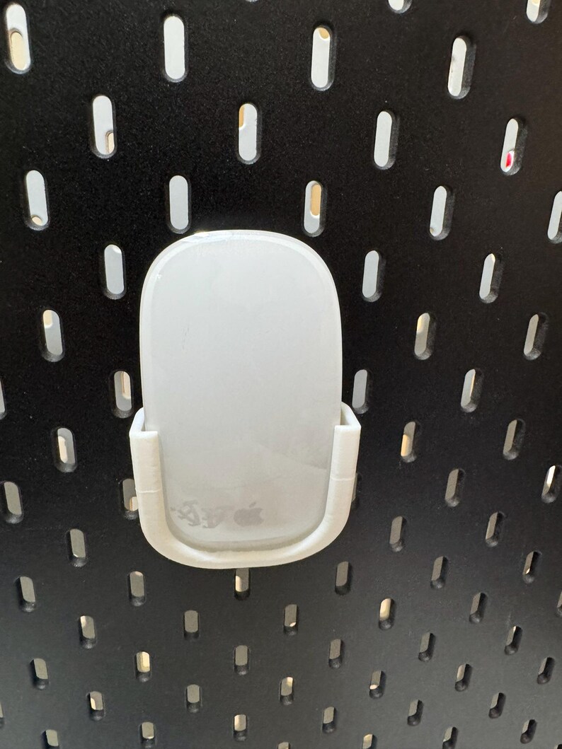 K&ouml;nnte beinhalten: Eine wei&szlig;e Computermaus wird in einer wei&szlig;en Kunststoffhalterung gehalten, die an einer schwarzen Lochwand befestigt ist. Die Maus hat ein glattes, abgerundetes Design und das Apple-Logo ist sichtbar. Die Lochwand hat gleichm&auml;&szlig;ig verteilte, ovale L&ouml;cher.