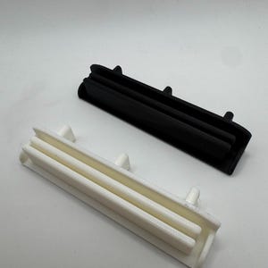 Pode incluir: Dois objetos retangulares de pl&aacute;stico, um preto e outro branco, s&atilde;o exibidos contra um fundo branco. Cada objeto tem um design canelado com pequenas protuber&acirc;ncias. Parecem ser componentes.