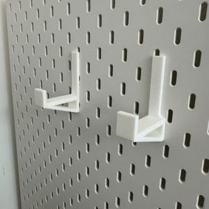 May include: A white pegboard with two white, L-shaped hooks attached. The pegboard has a grid of evenly spaced, oblong holes. The hooks are designed to fit into the holes, providing a storage or display solution. The pegboard is mounted on a wall.