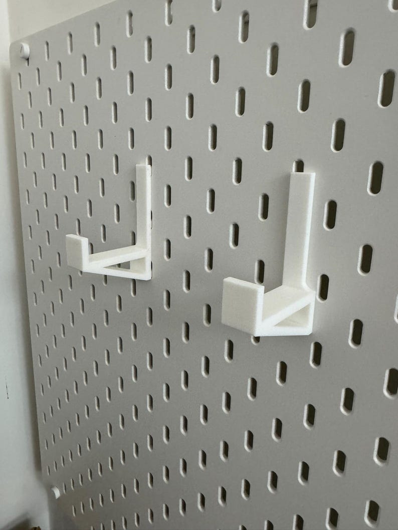 May include: A white pegboard with two white, L-shaped hooks attached. The pegboard has a grid of evenly spaced, oblong holes. The hooks are designed to fit into the holes, providing a storage or display solution. The pegboard is mounted on a wall.