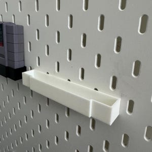 K&ouml;nnte beinhalten: Ein wei&szlig;es, rechteckiges Regal und eine graue Videospielkassette sind an einer wei&szlig;en Lochwand befestigt. Das Regal ist f&uuml;r kleine Gegenst&auml;nde konzipiert. Die Lochwand hat gleichm&auml;&szlig;ig verteilte, ovale L&ouml;cher.