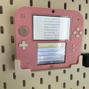 May include: A pink Nintendo 2DS handheld game console, mounted on a white pegboard. The screen displays a language selection menu with options in English, French, German, and Spanish. The console has a D-pad, buttons, and a stylus.