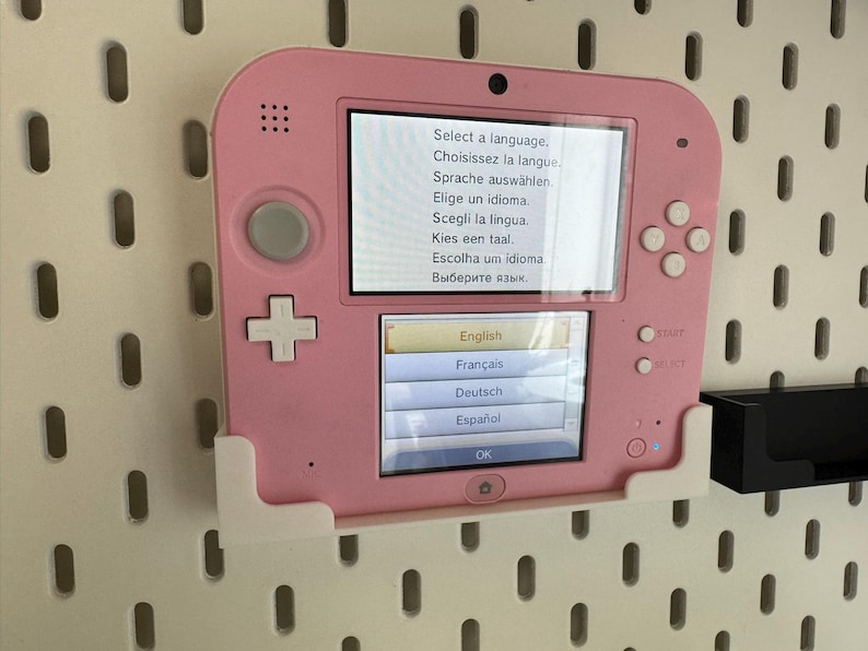 May include: A pink Nintendo 2DS handheld game console, mounted on a white pegboard. The screen displays a language selection menu with options in English, French, German, and Spanish. The console has a D-pad, buttons, and a stylus.