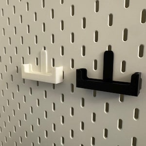 以下が含まれることがあります： 2つのペグボードアクセサリー：白と黒のホルダー。ホルダーは白いペグボードに取り付けられています。ホルダーは、垂直のサポートが付いたU字型のベースを持っています。