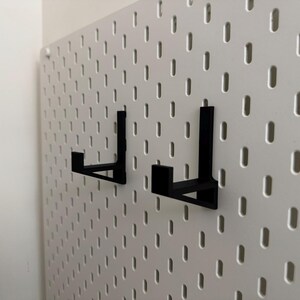 Op de afbeelding: Twee zwarte metalen pegboardhaken zijn gemonteerd op een witte pegboard. De haken zijn L-vormig en ontworpen om items vast te houden. Het pegboard heeft een raster van gelijkmatig verdeelde gaten.